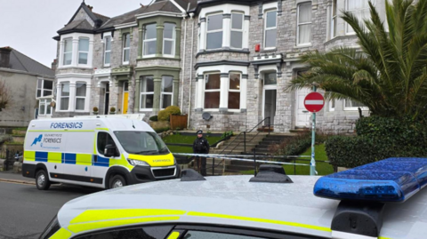 A police officer in uniform stands guard at a cordon outside a row of grey-bricked terraced houses in Plymouth. He is standing behind a line of police tape. A police forensics van is parked next to him on the road. The roof of a police car can also be seen.