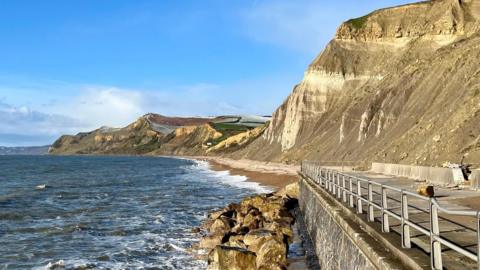 A cliffside view overlooking a beach. The sky is clear and blue with the stone cliffs towering over the beach and sea. There is no-one visible in the picture. A metal fence runs along a structure on the right of the picture.