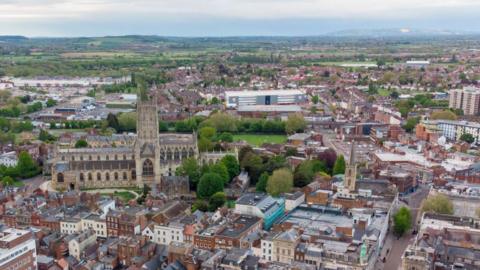 An aerial view of Gloucester city centre, which includes landmarks such as Gloucester Cathedral, Kingsholm Stadium, and the gate streets. The Cotswold hills can be seen in the distance.
