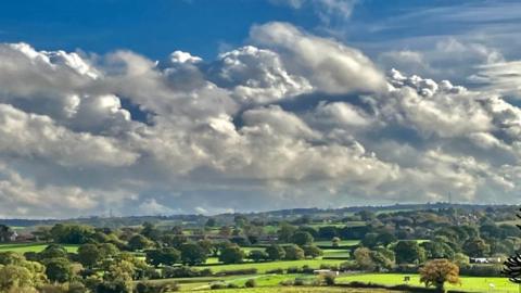 Fields and trees fill the lower half of the image and above the horizon there are lots of pillow-y white and grey clouds with a small amount of blue sky at the top.