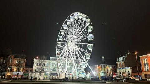 A white ferris wheel with a crane behind it and buildings in the background