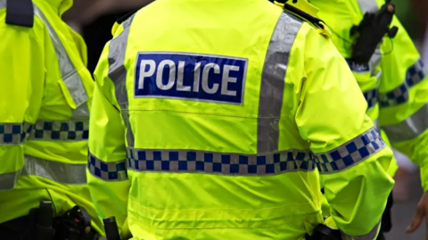 Image shows the back of a police officer's florescent jacket. There is a blue and white chequered band around it, and the word POLICE in white capital letters on a blue background.  