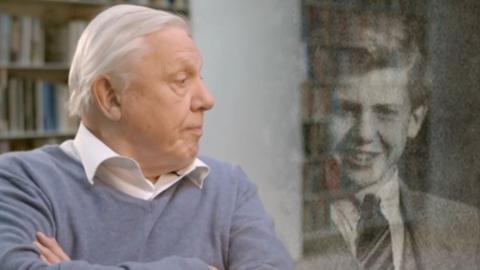 David Attenborough looks to his left, where there is a black and white picture of him as a child