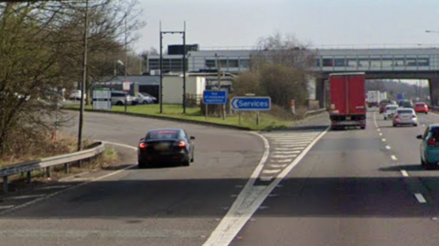 The slip road to a motorway services, with three lanes of traffic to the right of the services, and a covered bridge over the road