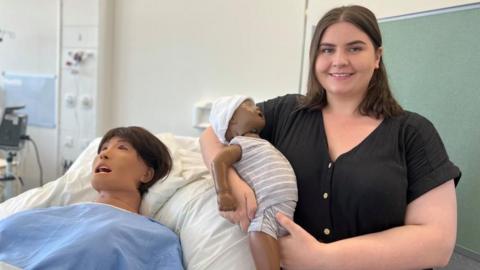 A lady wearing a black top is smiling holding a plastic baby wearing a grey striped babygrow. She is stood next to a hospital bed, with a dummy patient who symbolises a mother who has just given birth.