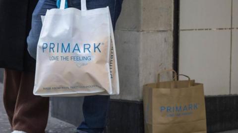 A shoppers leans against a wall outdoors holding a large white Primark bag while a smaller brown paper Primark bag sits on the floor