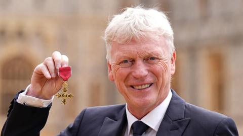 David Moyes stands in a suits smiling and holding up an OBE medal. 