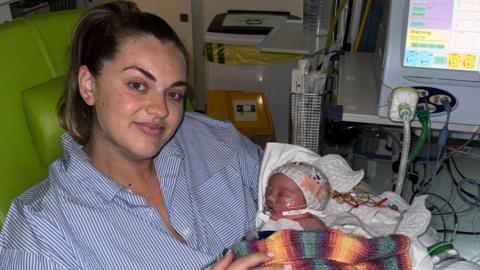 A woman with blonde hair, wearing a blue and white striped shirt, holds a newborn baby wrapped in a multicoloured blanket. There are tubes connecting the baby girl to a machine.