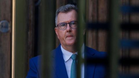Jeffrey Donaldson, a man in a blue suit with short grey hair and glasses, seen through the bars of an entrance gate.