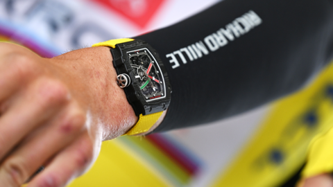 A close-up of a square watch with a black face and a yellow strap on the arm of a cyclist, who wears a Richard Mille sponsorship logo on his sleeve.