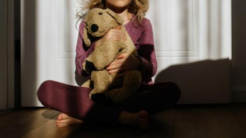 A stock image of an unidentified child holding a soft toy. They are sat on the floor with their back against a door.