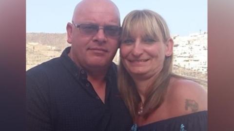 On the left, there is a man who is bald, wearing a black polo t-shirt with dark grey framed sunglasses. On the right, there is a woman in a dark blue off-the-shoulder t-shirt, with a tattoo next to her shoulder. She has blonde hair. 