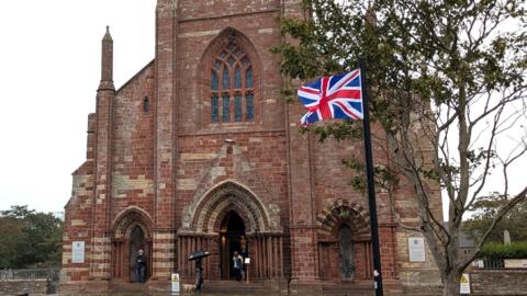 Brown brick cathedral with a large union jack flying on a pole in front of the building. There are people at the entrance of the cathedral and a tree in the foreground