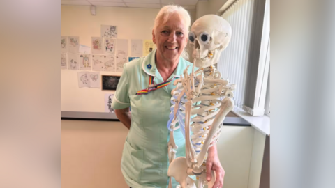 Sue Baker, who has been a nurse for 50 years, pictured beside a skeleton in green scrubs. Sue has white hair and is wearing a rainbow lanyard.