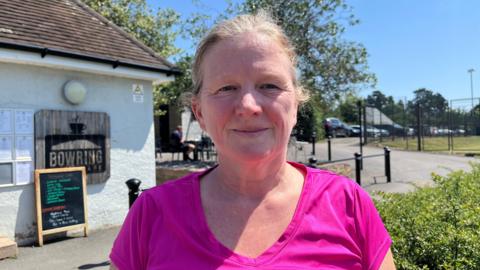 Donna pictured looking into the camera and smiling slightly. She has blonde grey hair tied up and is wearing a hot pink athletic top. She is standing outside the Bowring Park Cafe. It is a single-storey, small building, painted white and a chalkboard sign is standing outside it. 