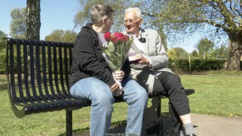 The photo shows a man and a woman sitting on a black metal bench. The woman is wearing a black hoodie and blue jeans. The man is wearing a grey blazer and black trousers. The woman is holding a bunch of red roses. They're sitting in a park. 