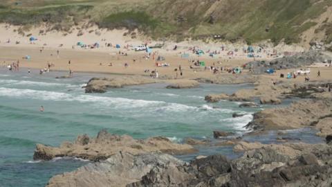 Fistral Beach is a wide sandy beach with lots of people walking and lying on it and there are people swimming in the sea alongside jagged rocks. There are many windbreaks and beach tents. It is a slightly cloudy day.