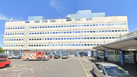 The long, grey concrete building is six storeys tall and has blue blocks on the top. There is a fire engine parked out front in the car park which is also dotted with cars. A smaller building on the right of the frame has a: "Welcome to University Hospital of North Tees" sign.