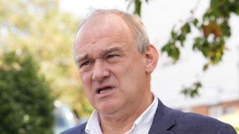 Sir Ed Davey has open collar and black suit on. He is looking afar. He has grey hair on the sides of his head, which is thin on top.