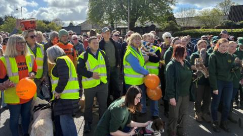 A crowd of people gathered in a town - some are wearing hi-vis jackets, others have orange shirts which say "Save Eccleshall".