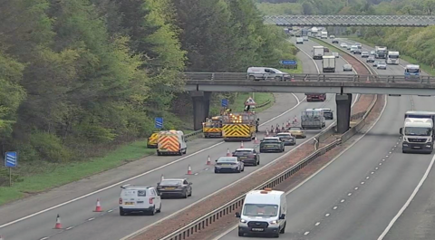 A motorway, with one carriageway blocked off by cones, and repair vehicles underneath a bridge.