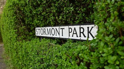 A street sign - black font on a white background - reading "Stormont Park". It is completely surrounded by a green garden hedge.