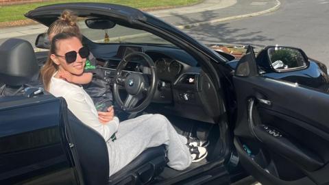 Hannah Taylor in BMW with child on lap. The door is open and she is wearing sunglasses. 