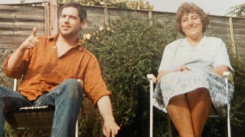 Nigel Goodman is sitting on a deck chair to the left, wearing an oversized orange t-shirt and blue denim jeans. To his right, on a deck chair, is his mother Heather, wearing a shirt and skirt.