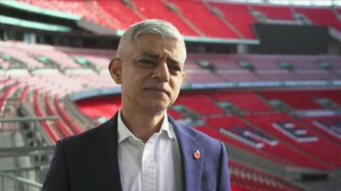 Sadiq Khan stood in the stands at Wembley Stadium. He is wearing a white shirt with an unbuttoned collar and a blue jacket, with a straight face looking at the camera.