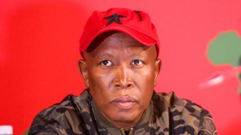 Leader of the South African opposition party Julius Malema wearing a red cap, with a black star and a camouflage jacket. He looks serious.