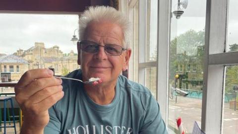 David Earley pictured in a cafe with a spoonful of cake in hand, wearing a light blue Hollister t-shirt and wearing wire rim glasses