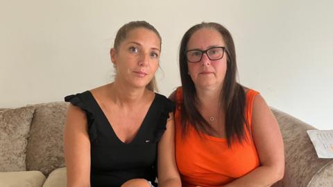 Two women look at the camera. Both have long dark hair. One is wearing an orange top and the other is wearing black. 