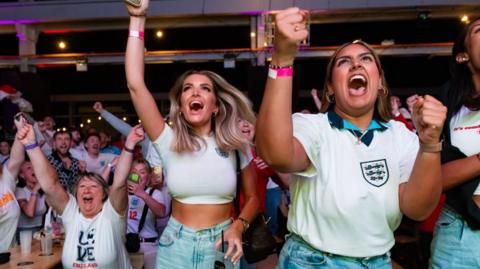 Fans in England football shirts celebrate a goal in a bar. Closest to the camera are two women in white England branded tops and jeans. Behind them and to their right an older woman can be seen celebrating while sat down. Dozens of other fans are in the background.