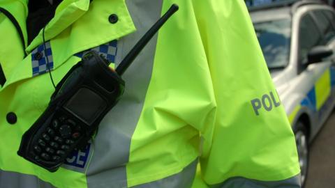 Close-up of fluorescent yellow police jacket with a black walkie-talkie attached. There is a police car in the background