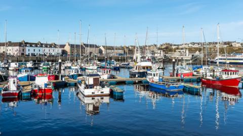 Wick harbour with different types of colourful boats tied up next to pontoons. Along the shore are traditional, three-storey buildings. The sky above is blue and cloudless.
