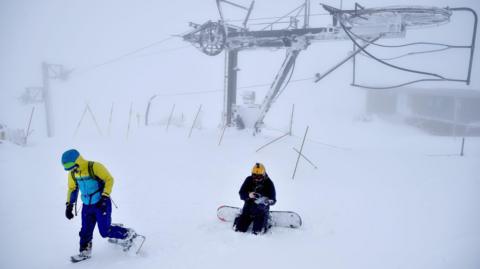 Two snowboarders in whiteout conditions at Cairngorm Mountain. There is ski lift infrastructure in the background.