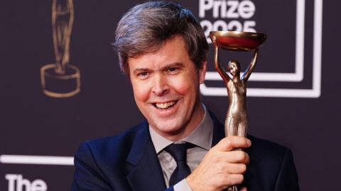 David Szalay smiling and holding up the Booker Prize trophy