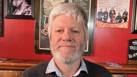 Jason O'Rourke has short white hair and a white and grey beard. He is sitting in a bar with old signs behind him. He is wearing a navy jumper and light blue patterned shirt underneath.