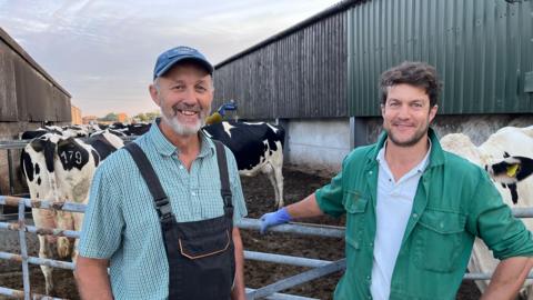 Rob and Will Warnock lean against a metal gate in the farmyard at Tumble Tye Farm in Capel-le-Ferne with a pen full of cows in the background