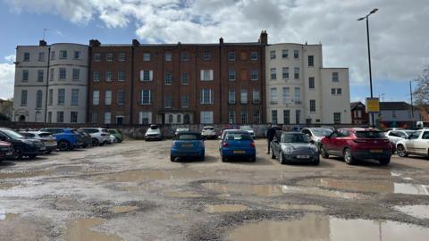 A car park, which has large puddles due to uneven surfacing, is overlooked by large but rundown townhouses in Cheltenham. There are plenty of cars parked in the car park.