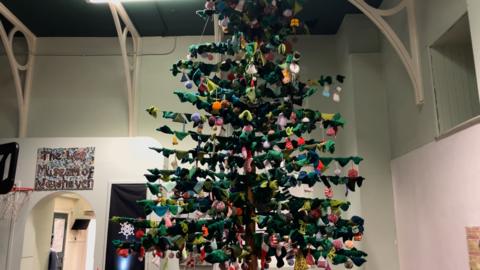 A 16ft knitted Christmas tree. The trunk of the tree is made of wood with wooden pegs sticking out from it. Knitted squares in colours of green are attached to look like branches. Other knitted decorations like stockings and baubles are attached.