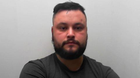 A mugshot of Russell, who is standing against a plain light-coloured wall. He is wearing a dark short-sleeved t-shirt and has both arms crossed in front of the chest. He has brown hair and a beard.