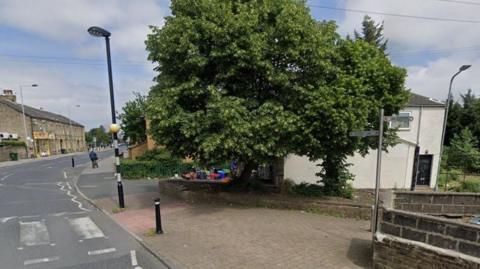 A large leafed tree stands on a raised platform next to a pavement. A zebra crossing is to the left of the picture, while a signpost is in the foreground.