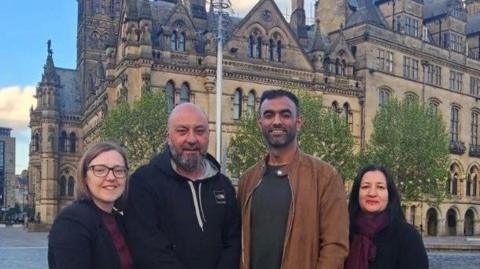 Two men and two women stand in a huddle in front of Bradford City Hall