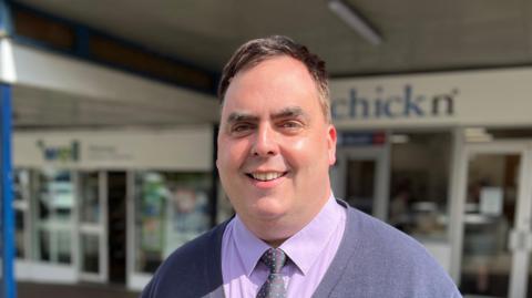 A smiling Kerry Smith looks directly at the camera as he stands outside a parade of shops