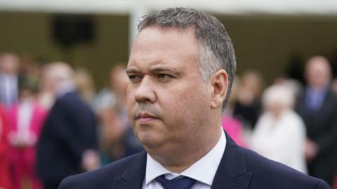 John Caldwell, with short grey hair, wearing a white shirt, navy tie and navy suit. He is pictured from the shoulders up. The background is blurred. 