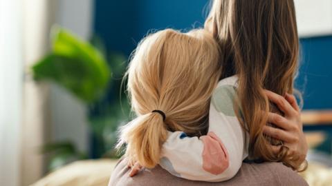 A mother and daughter embrace in their home with their backs to the viewer.