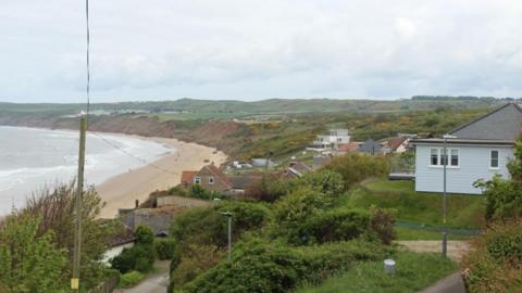 A number of cliff-top homes close to an eroded green slope leading to a stretch of sand and sea