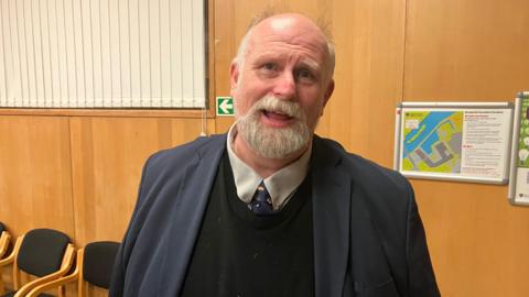 Tom Wootton is looking into the camera while standing in a wood-panelled room that appears to be an office or meeting space. He has white hair on the sides of his head and a beard. He is dressed in a dark blue suit jacket over a black sweater, with a white collared shirt and patterned tie. Behind him are wall-mounted evacuation diagrams and a row of black cushioned chairs with wooden frames.