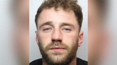 Police custody image of James Saxon with brown beard and hair wearing a black t-shirt.